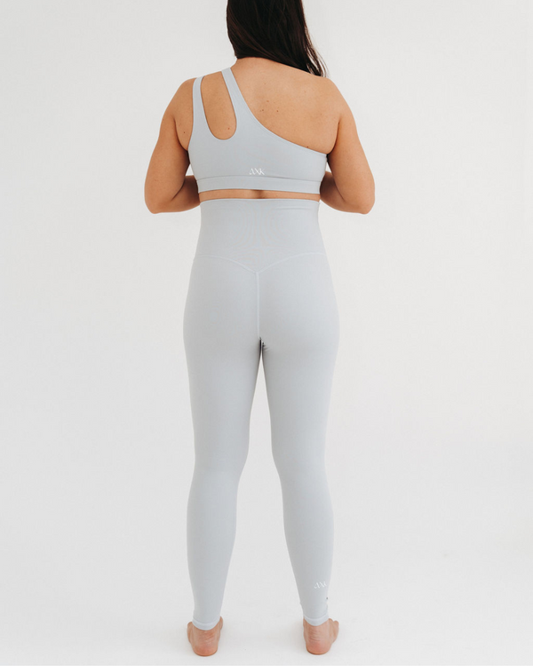 Back view of a pregnant woman wearing premium light grey maternity leggings with a flattering V-seam back and matching one-shoulder, low-impact, maternity and nursing sports bra, standing against a white background. Premium maternity athletic and activewear by AXK.