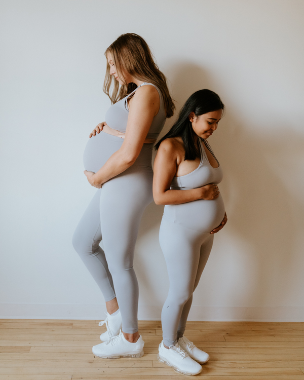 Two pregnant women wearing premium light grey maternity leggings with a flattering scrunch butt and matching one-shoulder, low-impact, nursing sports bras, standing side by side on a wood floor against a white wall. Premium maternity athletic and activewear by AXK.