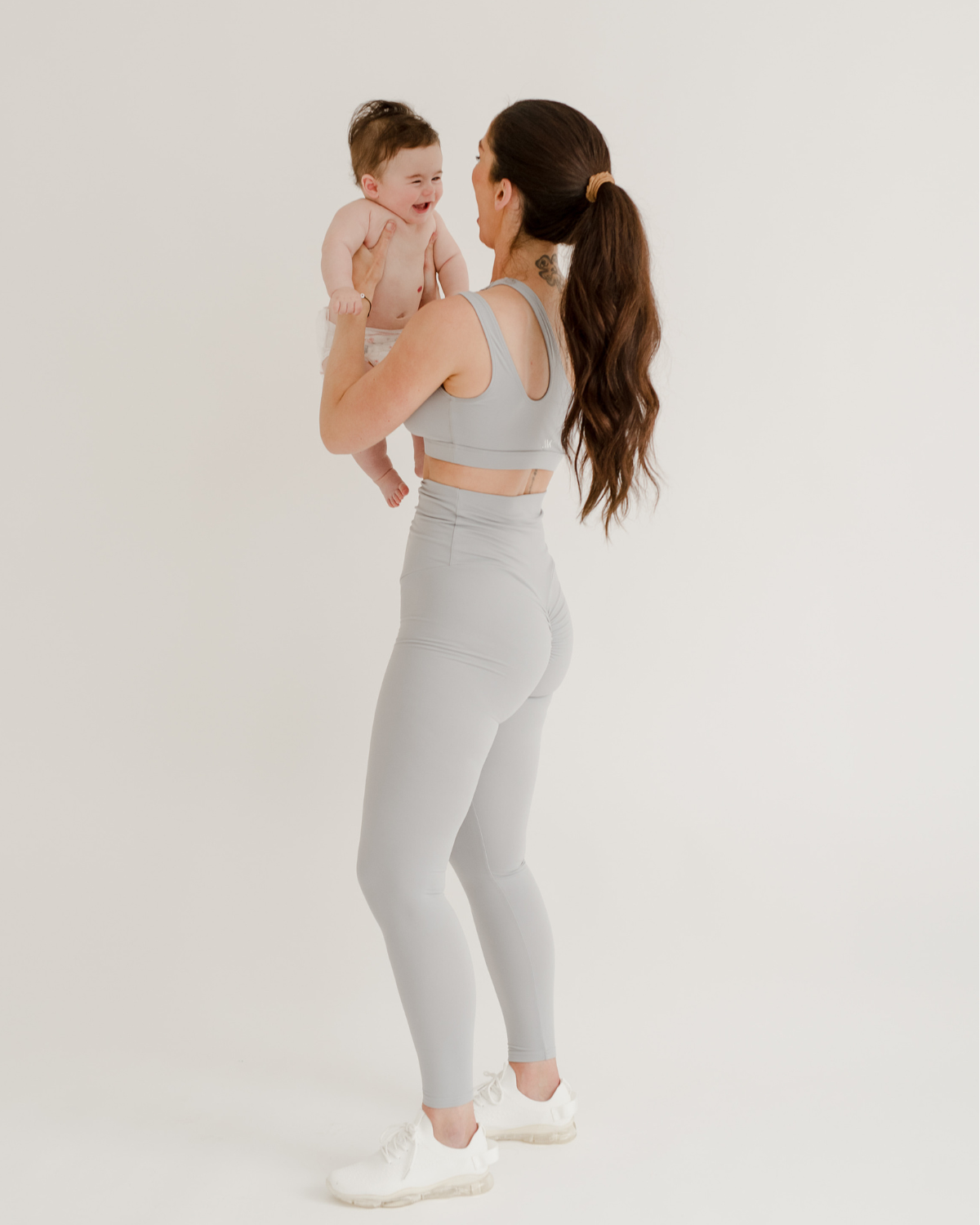 Mother wearing premium light grey maternity leggings with a flattering scrunch butt and matching, one-shoulder, low-impact, nursing sports bra, holding her smiling baby against a white background. Premium maternity athletic and activewear by AXK.