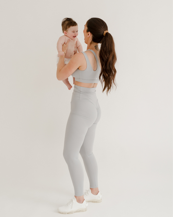 Mother wearing premium light grey maternity leggings with a flattering scrunch butt and matching, one-shoulder, low-impact, nursing sports bra, holding her smiling baby against a white background. Premium maternity athletic and activewear by AXK.