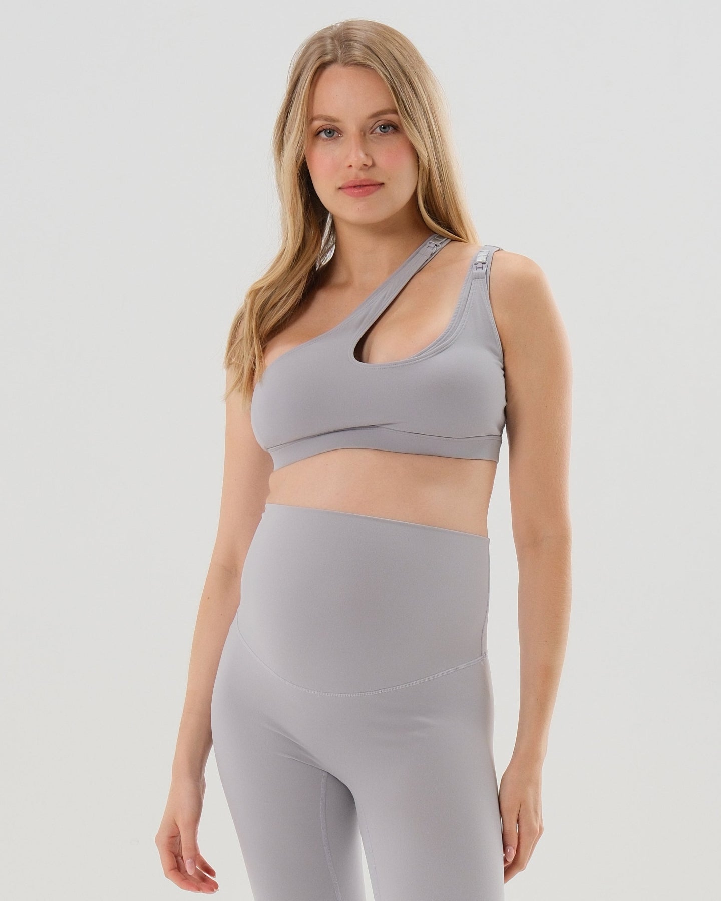 Pregnant woman in a one-shoulder light gray maternity and nursing sports bra, wearing matching light gray maternity leggings. Best selling 2026 premium maternity athletic and activewear by AXK, offered in regular and plus sizes, with glowing 5 star reviews. 