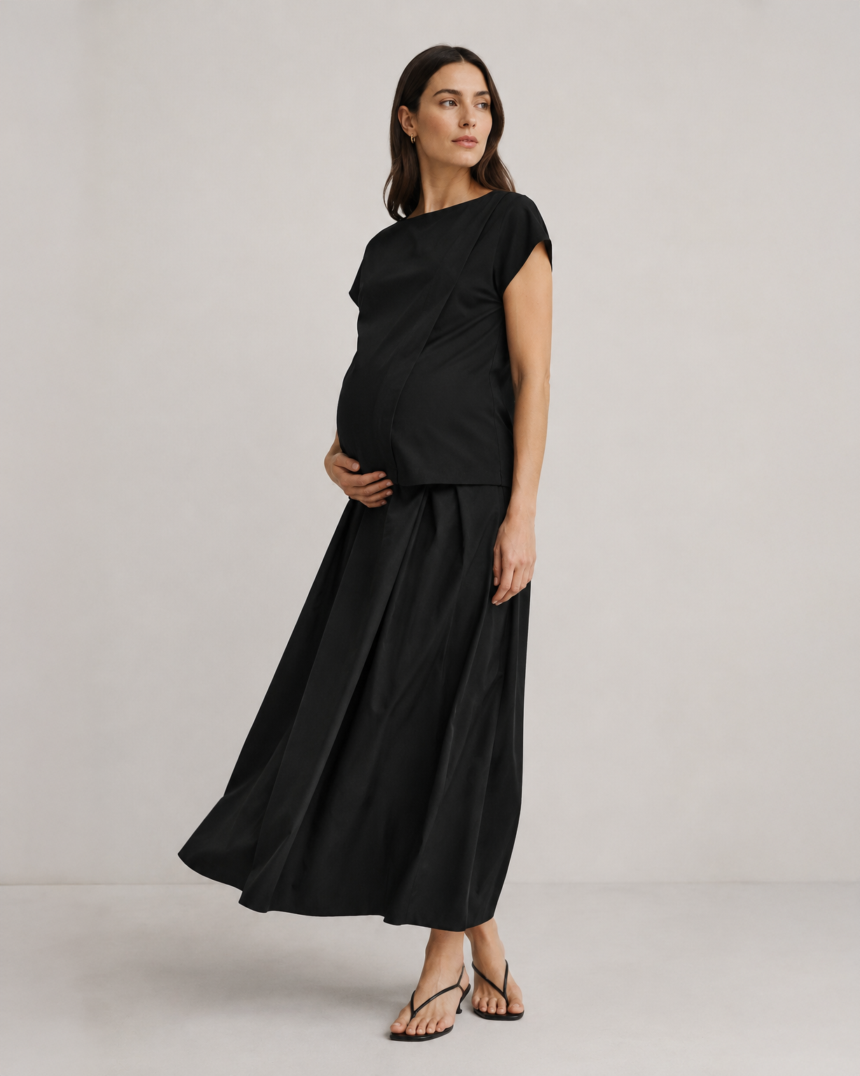 Pregnant woman wearing a black pleated designer maternity maxi maternity skirt in lightweight tropical wool by MARION. This luxury skirt can be worn over or under your belly bump. It fastens with a zip, and features deep functional pockets and a flexible elastic waistband for comfort. This refined, timeless style epitomizes quiet luxury. Perfect for maternity work wear, wedding guest dress, occasion wear, pregnancy street style, and baby shower outfits. Petite and tall friendly.