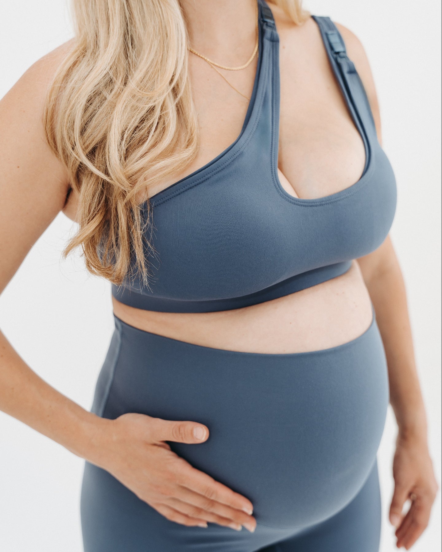 Pregnant woman in a one-shoulder blue maternity and nursing sports bra, wearing matching blue maternity leggings. Best selling 2026 premium maternity athletic and activewear by AXK, offered in regular and plus sizes, with glowing 5 star reviews. 