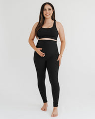 Pregnant woman wearing a black maternity set, including black maternity leggings with a flattering scrunch butt and a one-shoulder black maternity and nursing sports bra, on a white background.