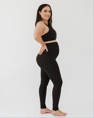 Pregnant woman wearing a black maternity set, including black maternity leggings with a flattering scrunch butt and a one-shoulder black maternity and nursing sports bra, on a white background.