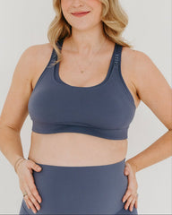 Pregnant woman wearing a blue maternity and nursing sports bra and pregnancy leggings on a plain background.