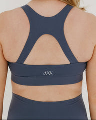 Pregnant woman wearing a blue maternity and nursing sports bra and pregnancy leggings on a plain background.