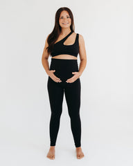 Pregnant woman wearing a black maternity set, including black maternity leggings and a one-shoulder black maternity and nursing sports bra, on a white background.