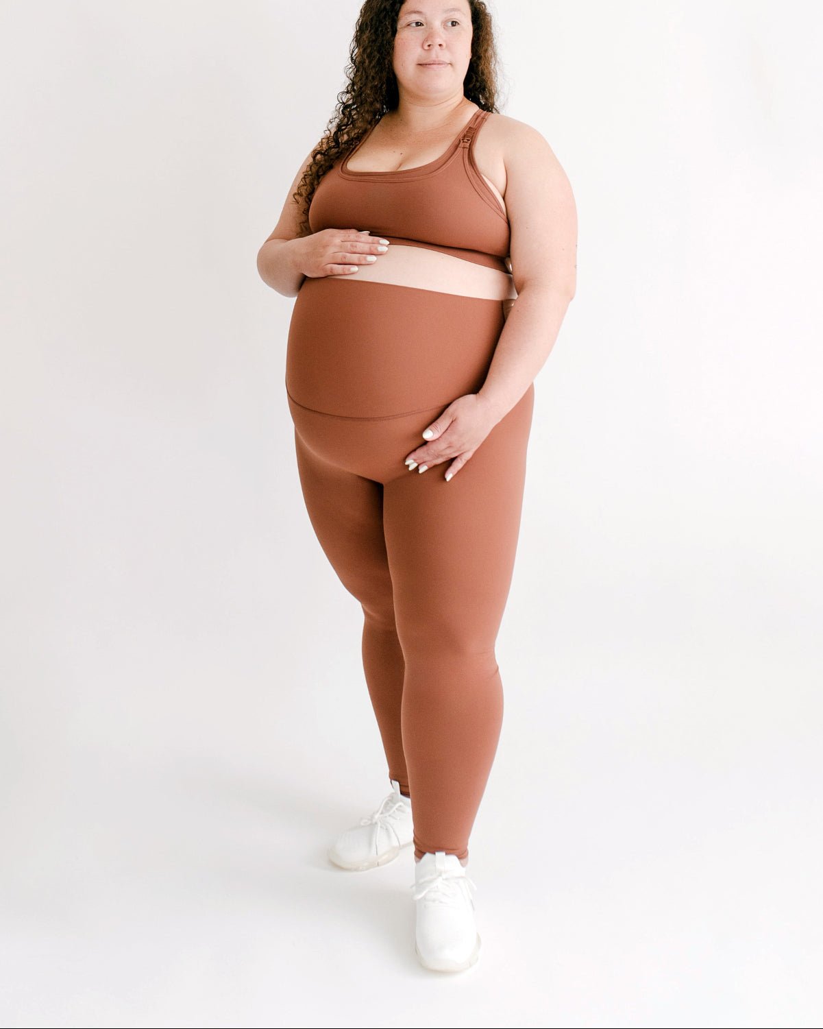 Woman wearing a brown maternity set, including one-shoulder brown maternity and nursing sports bra and brown maternity leggings, on a white background.