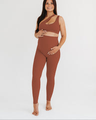 Woman wearing a brown maternity set, including one-shoulder brown maternity and nursing sports bra and brown maternity leggings, on a white background.