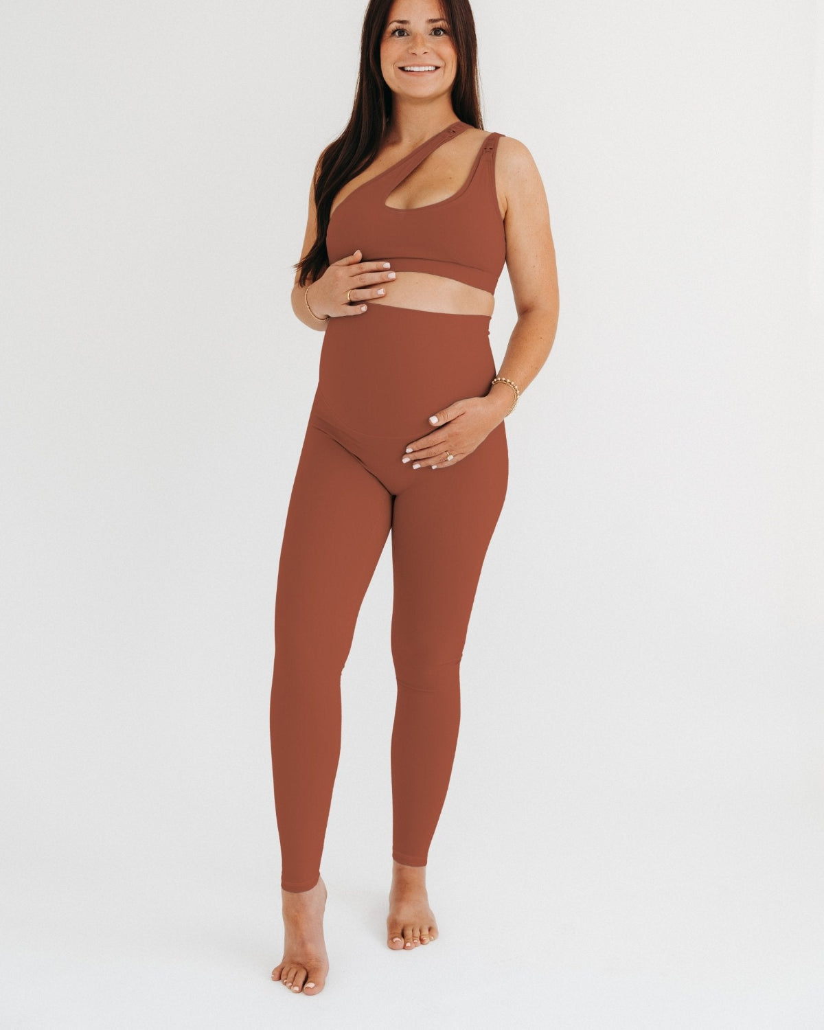 Pregnant woman wearing premium brown AXK maternity leggings with a supportive high waist and matching high-impact racerback maternity and nursing sports bra, standing against a white background. Premium maternity athletic and activewear by AXK - high quality petite, tall, and plus size maternity. 