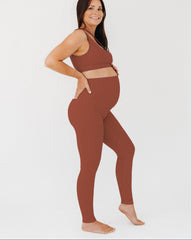 Woman wearing a brown maternity set, including one-shoulder brown maternity and nursing sports bra and brown maternity leggings, on a white background.