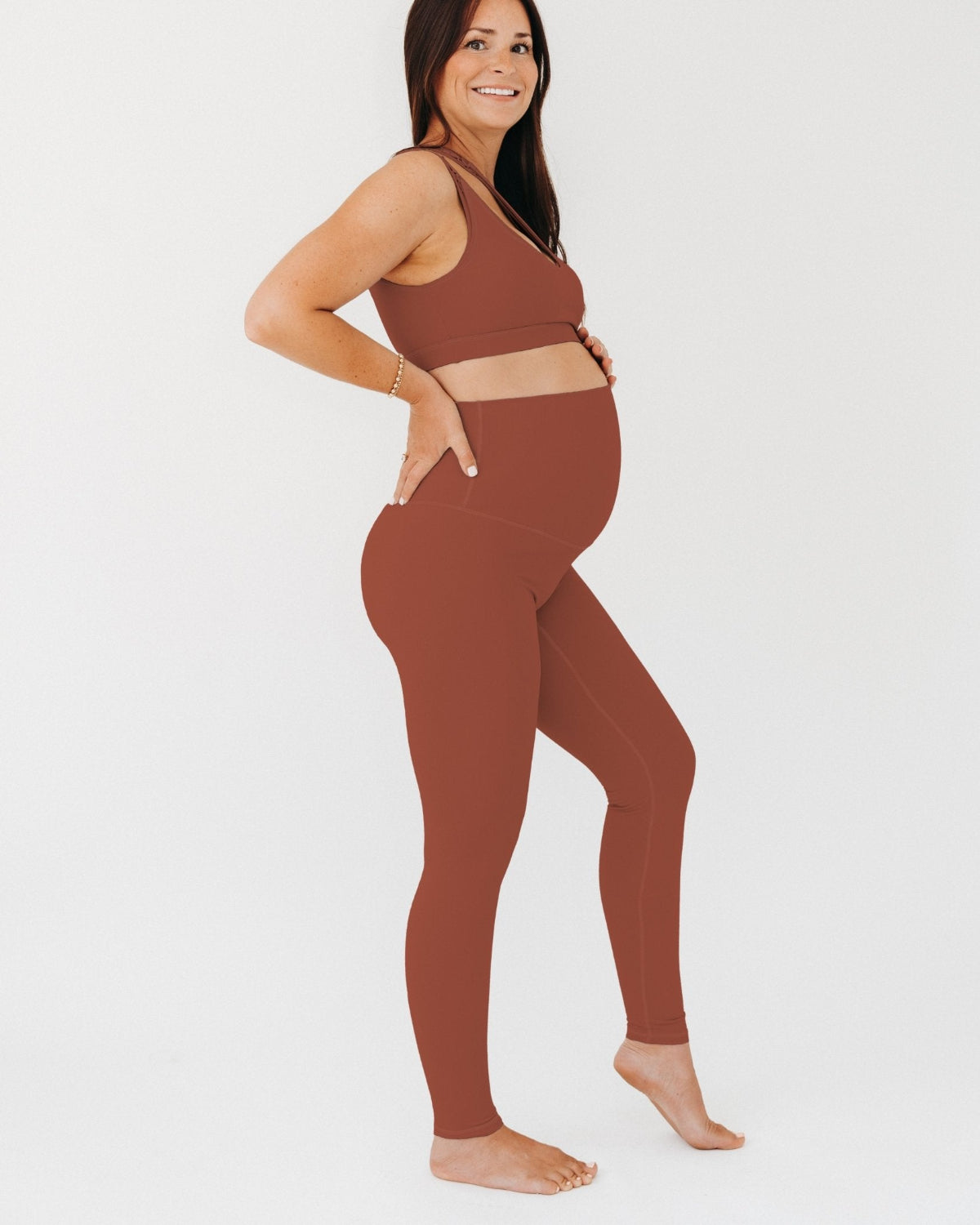 Pregnant woman wearing premium brown AXK maternity leggings with a supportive high waist and matching high-impact racerback maternity and nursing sports bra, standing against a white background. Premium maternity athletic and activewear by AXK - high quality petite, tall, and plus size maternity. 