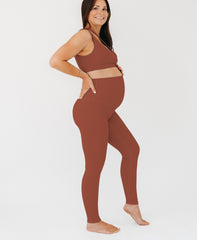 Pregnant woman wearing premium brown AXK maternity leggings with a supportive high waist and matching high-impact racerback maternity and nursing sports bra, standing against a white background. Premium maternity athletic and activewear by AXK - high quality petite, tall, and plus size maternity. 