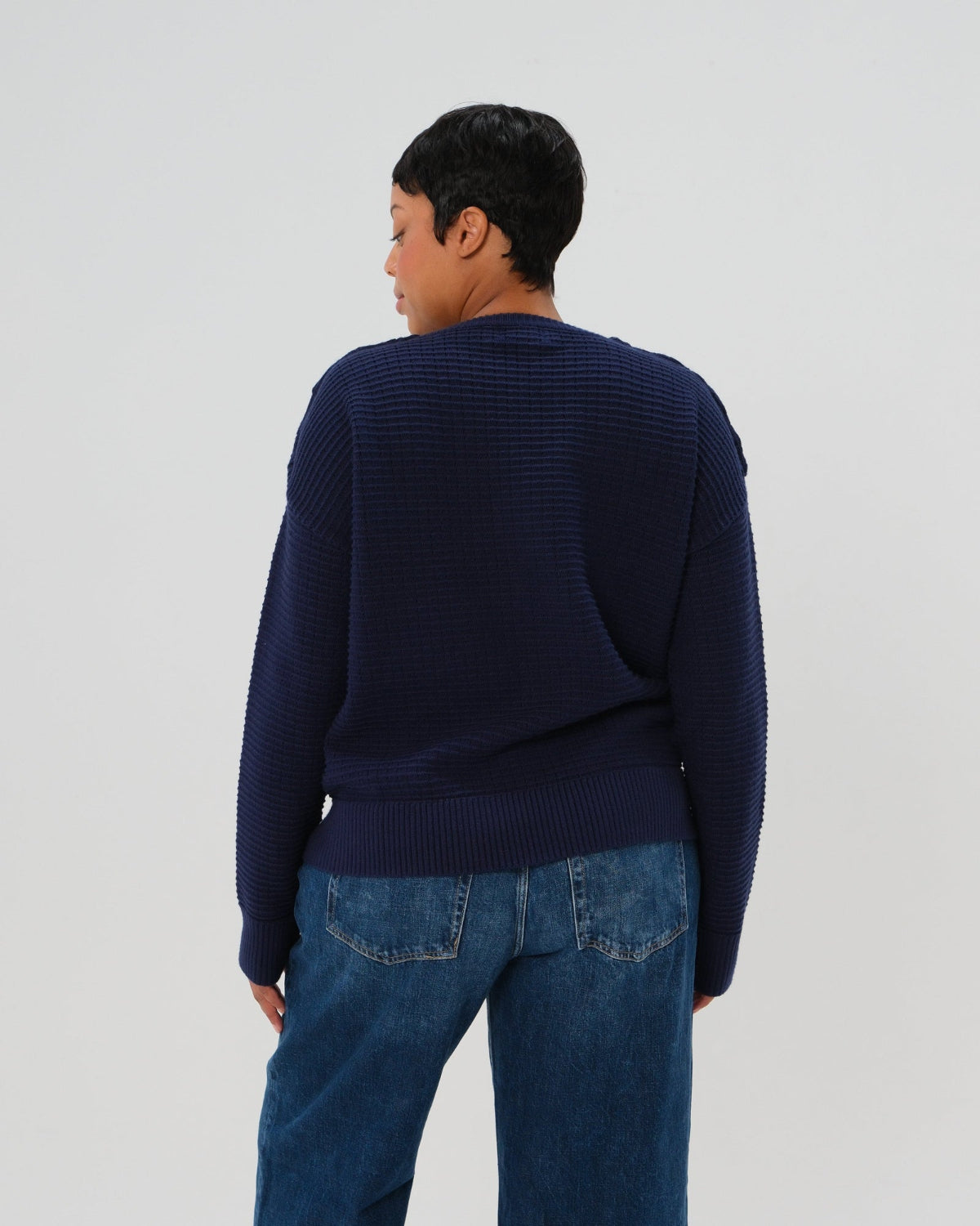 Navy premium maternity and nursing sweater in stretch cotton box knit. Featuring shoulder button detail for breastfeeding and discreet hem thumbholes for an extra cozy fit. Oversized fit is flattering, polished, and versatile - transitioning from pregnancy workwear to loungewear to streetwear with ease. Petite and tall friendly; available in regular and plus size. Back view.