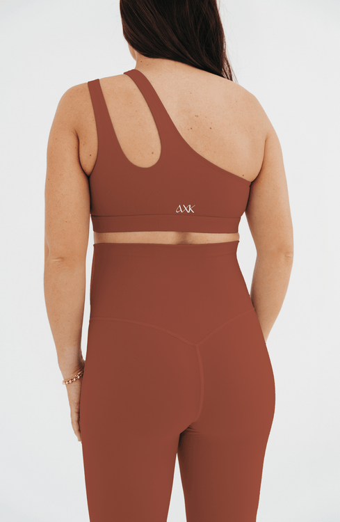 Back view of a woman wearing a brown one-shoulder maternity and nursing sports bra with matching high-waist leggings featuring a V-seam back, standing against a white background. Premium maternity activewear by AXK.