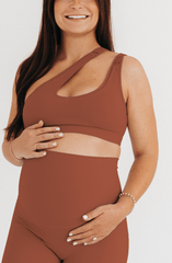 Pregnant woman wearing a brown one-shoulder maternity and nursing sports bra with matching high-waist leggings, standing against a white background. Premium maternity activewear by AXK.