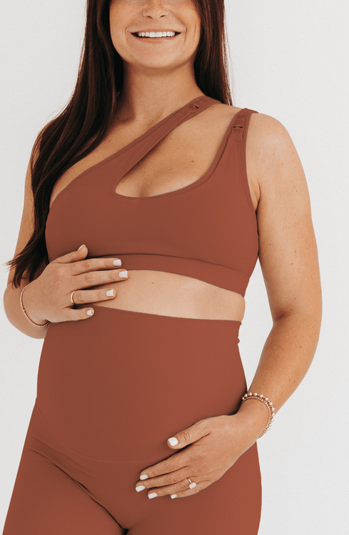 Pregnant woman wearing a brown one-shoulder maternity and nursing sports bra with matching high-waist leggings, standing against a white background. Premium maternity activewear by AXK.