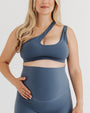 Pregnant woman wearing premium maternity leggings with a supportive high waist and a matching one-shoulder maternity and nursing sports bra in blue, standing against a white background. Premium maternity athletic and activewear by AXK.