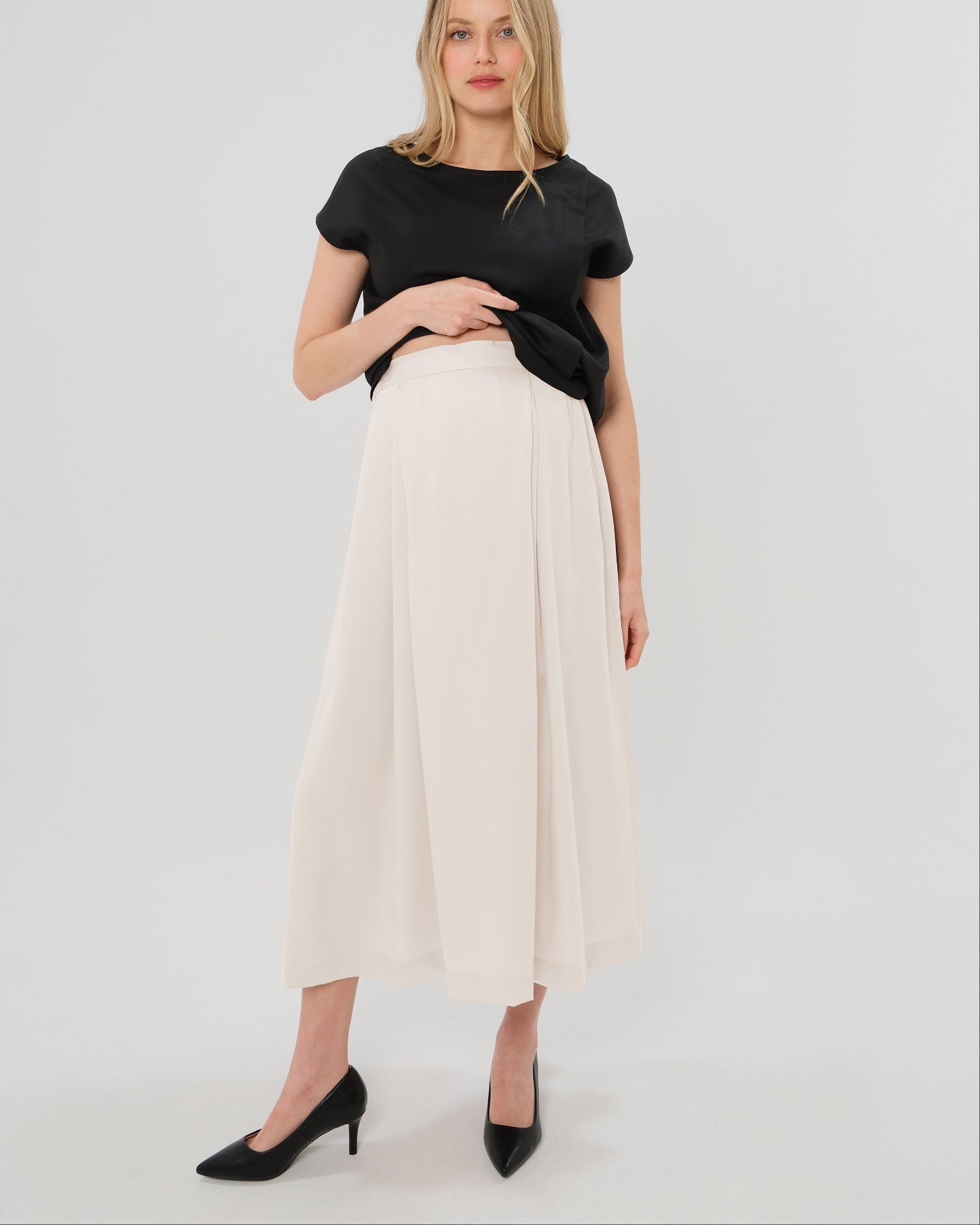 Pregnant woman wearing a modest off-white cream pleated designer maternity maxi maternity skirt in luxurious TENCEL satin by MARION. This luxury skirt can be worn over or under your belly bump. It fastens with a zip, and features deep functional pockets and a flexible elastic waistband for comfort. This refined, timeless style epitomizes quiet luxury. Perfect for maternity work wear, wedding guest dress, occasion wear, pregnancy street style, and baby shower outfits. Petite and tall friendly.