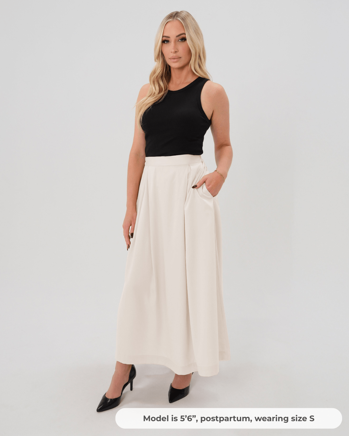 Postpartum woman wearing a modest off-white cream pleated designer maternity maxi maternity skirt in luxurious TENCEL satin by MARION. This luxury skirt can be worn over or under your belly bump. It fastens with a zip, and features deep functional pockets and a flexible elastic waistband for comfort. This refined, timeless style epitomizes quiet luxury. Perfect for maternity work wear, wedding guest dress, occasion wear, pregnancy street style, and baby shower outfits. Petite and tall friendly.