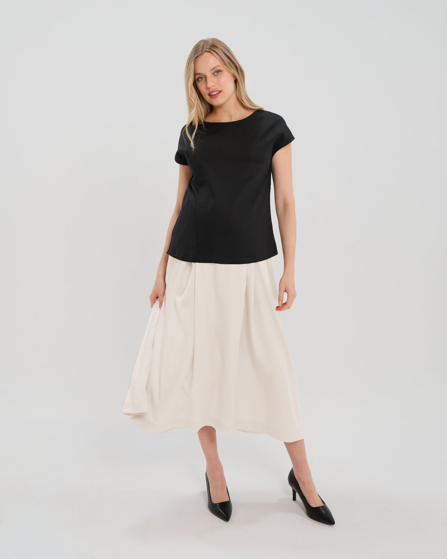 Pregnant woman wearing a modest off-white cream pleated designer maternity maxi maternity skirt in luxurious TENCEL satin by MARION. This luxury skirt can be worn over or under your belly bump. It fastens with a zip, and features deep functional pockets and a flexible elastic waistband for comfort. This refined, timeless style epitomizes quiet luxury. Perfect for maternity work wear, wedding guest dress, occasion wear, pregnancy street style, and baby shower outfits. Petite and tall friendly.