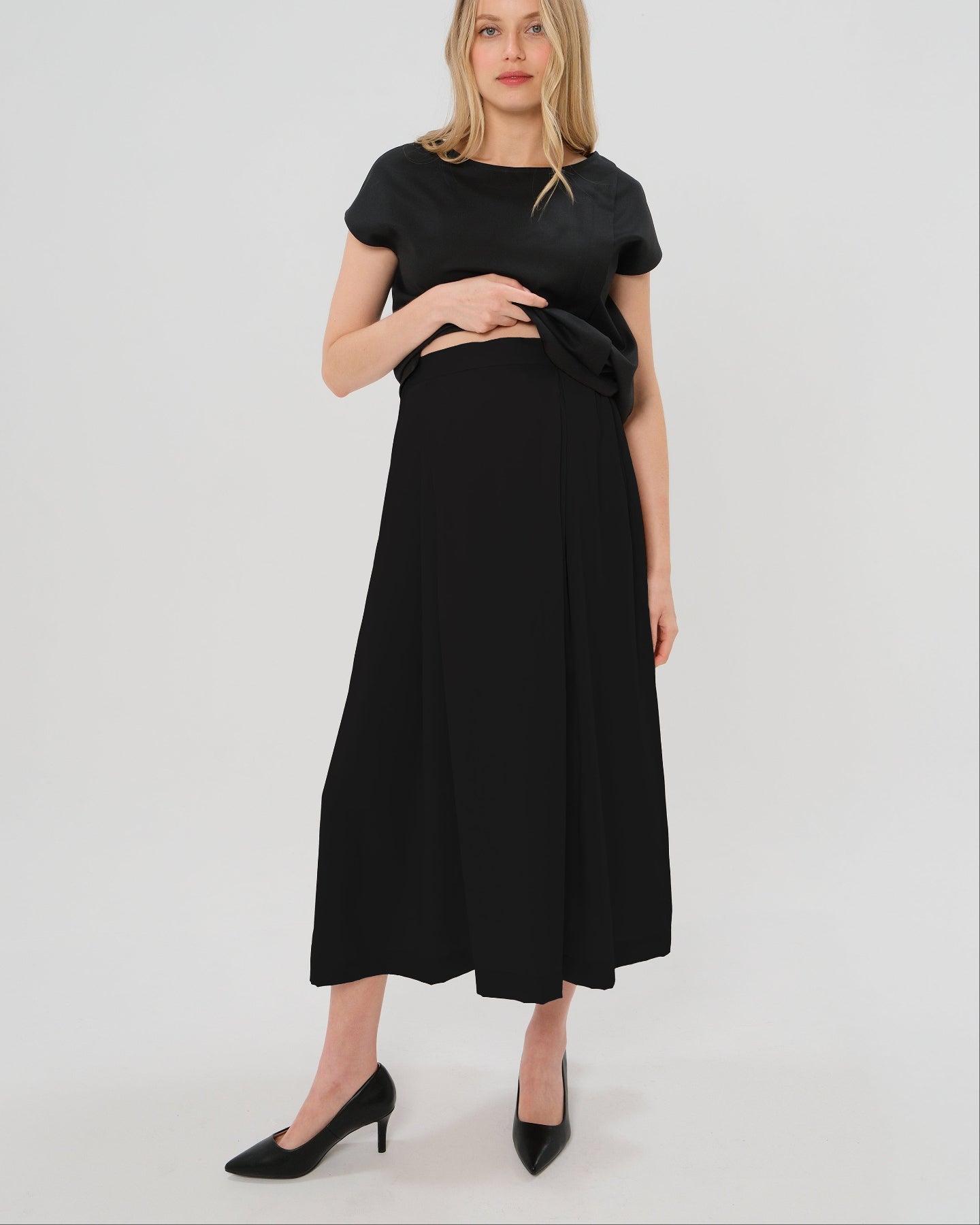 Pregnant woman wearing a black pleated designer maternity maxi maternity skirt in lightweight tropical wool by MARION. This luxury skirt can be worn over or under your belly bump. It fastens with a zip, and features deep functional pockets and a flexible elastic waistband for comfort. This refined, timeless style epitomizes quiet luxury. Perfect for maternity work wear, wedding guest dress, occasion wear, pregnancy street style, and baby shower outfits. Petite and tall friendly.