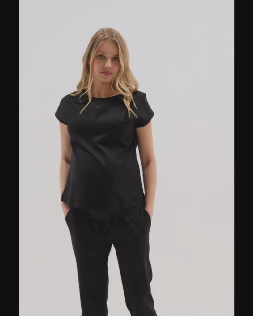 Video of a pregnant woman wearing a black maternity short sleeve boat neck shell top with designer nursing zipper and easy, flattering elegance for work or going out. Cut in breathable, all-season tropical wool blend. This bestselling business apparel item features cap sleeves perfect for spring and summer pregnancy or breastfeeding; transitions perfectly for layering under jackets or sweaters in fall and winter. 